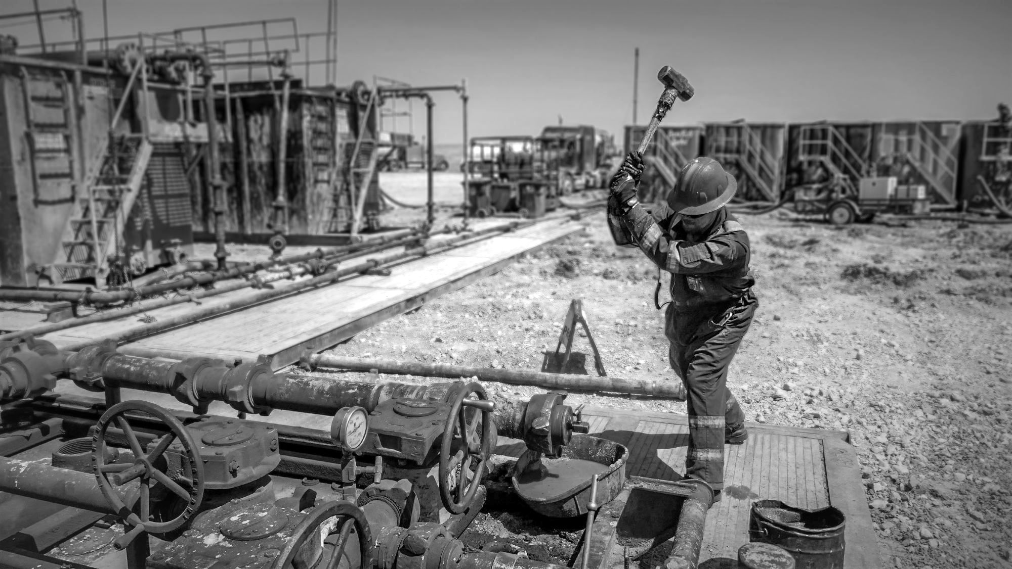 Oilfield hand in a hard hat swinging a sledgehammer on a frac site